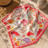 Pañoleta satinada Coralina de 70 × 70 cm con estampado tropical en tonos coral, rosa y marfil, presentada junto a un sombrero de fibra natural en un escenario editorial de verano.