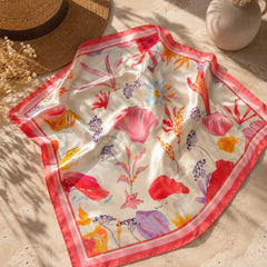 Pañoleta satinada Coralina de 70 × 70 cm con estampado tropical en tonos coral, rosa y marfil, presentada junto a un sombrero de fibra natural en un escenario editorial de verano.