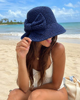 Modelo en la playa usando el sombrero bucket color azul marino tejido de papel reciclado con traje de baño blanco