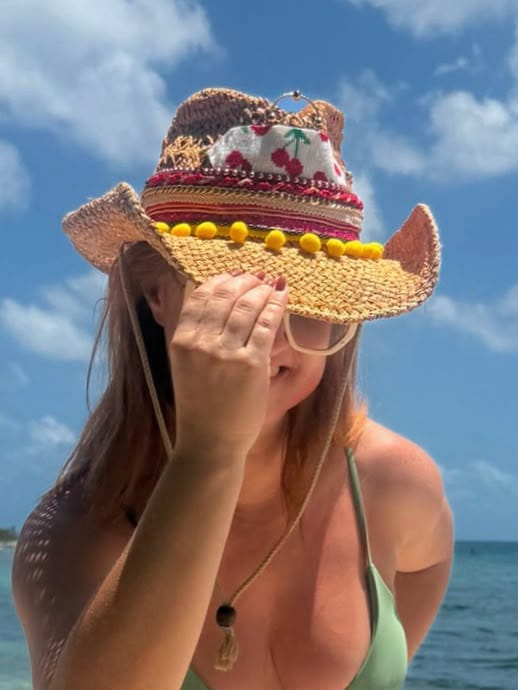 Modelo luciendo el sombrero Amore Cereza estilo vaquero con bordado de cerezas y pompones amarillos en un look de playa de verano.