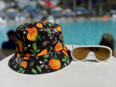 Sombrero Bucket Doble Vista Citrus con estampado de cítricos junto a gafas de sol blancas en un ambiente de piscina tropical.