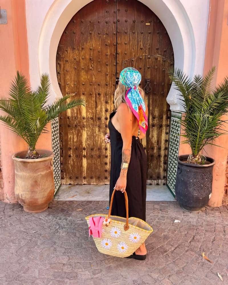Mujer luciendo pañoleta grande de seda Isla Fiesta anudada en la cabeza con vestido negro y bolso de playa con diseño de flores.