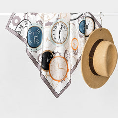 Pañoleta satinada Le Temps de 70 × 70 cm con ilustración de relojes clásicos junto a un sombrero de fibra natural, inspirando un estilo elegante y atemporal.