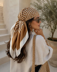 Modelo con pañoleta satinada Riviera Camel de 90 × 90 cm anudada en el cabello, combinada con bolso tejido y look de inspiración mediterránea.